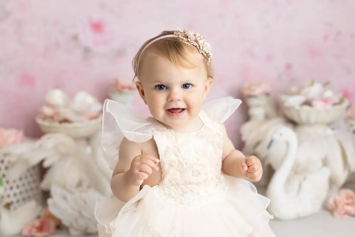 A baby wearing a white lace dress and a floral headband stands in front of a pastel pink background with decorative white swans and flowers. Lovely Baby Photography by Stacey Marsh