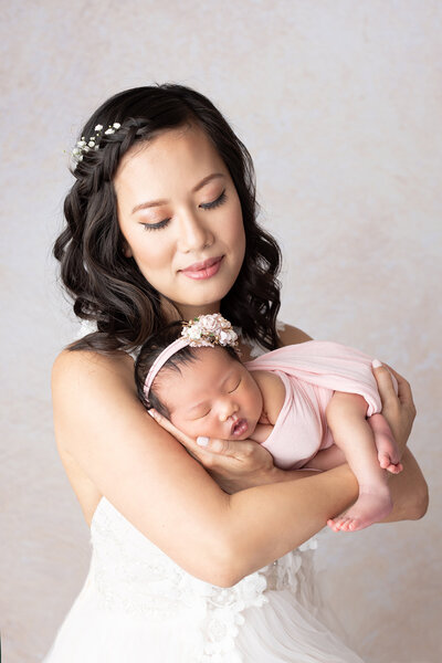 A woman in a white dress holds a sleeping newborn dressed in pink, both facing the camera against a light, neutral background. Lovely Baby Photography by Stacey Marsh