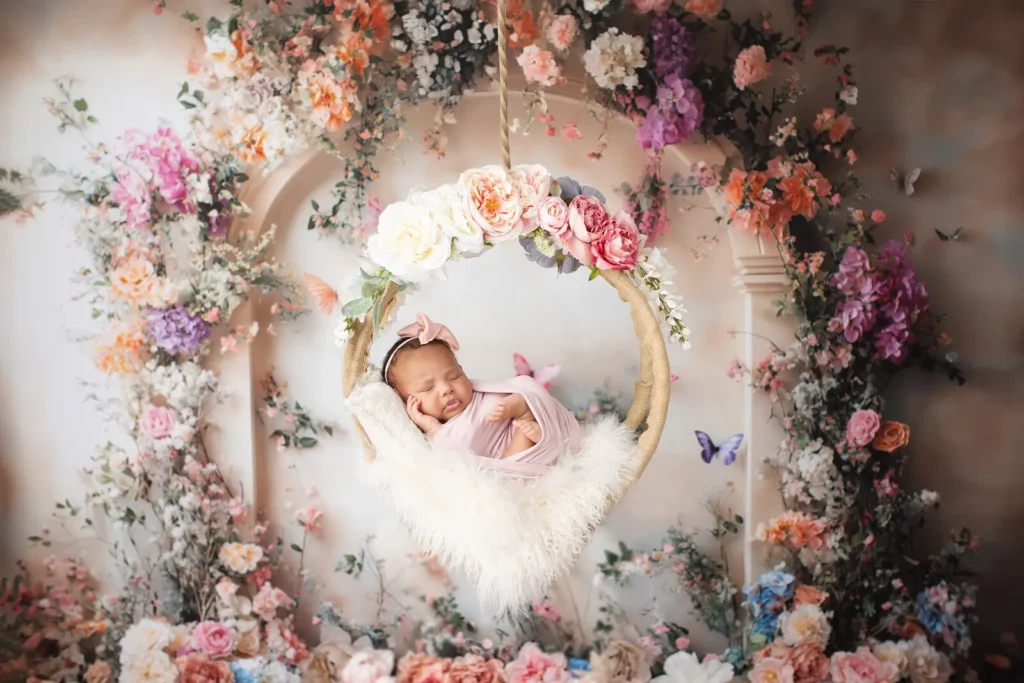 A newborn baby in a pink outfit and headband sleeps on a fur-lined swing, surrounded by colorful artificial flowers and butterflies. Lovely Baby Photography by Stacey Marsh