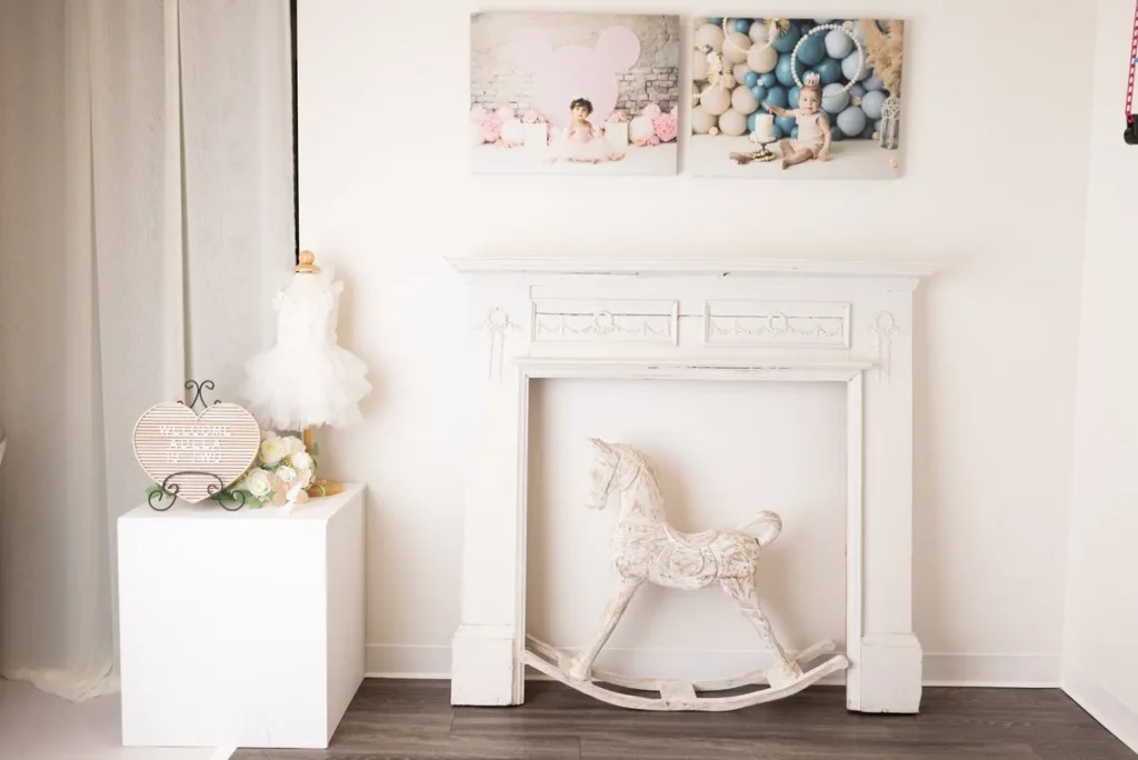 A white faux fireplace with a decorative rocking horse, a child’s dress on display, and two baby portraits above in a softly lit, minimalist room. Lovely Baby Photography by Stacey Marsh