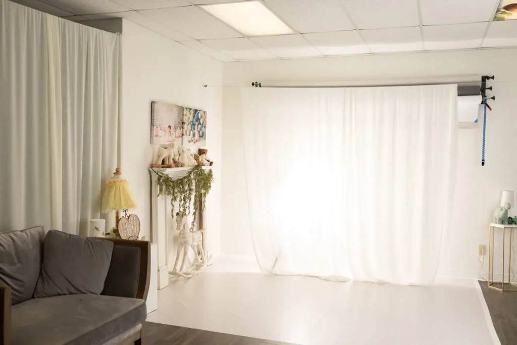 A photography studio setup with a white backdrop, neutral decor, a gray sofa, and a soft light illuminating the scene. Lovely Baby Photography by Stacey Marsh
