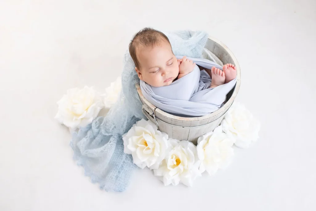 A sleeping baby wrapped in a light blue blanket is nestled inside a wooden bucket, surrounded by white artificial flowers on a white background. Lovely Baby Photography by Stacey Marsh