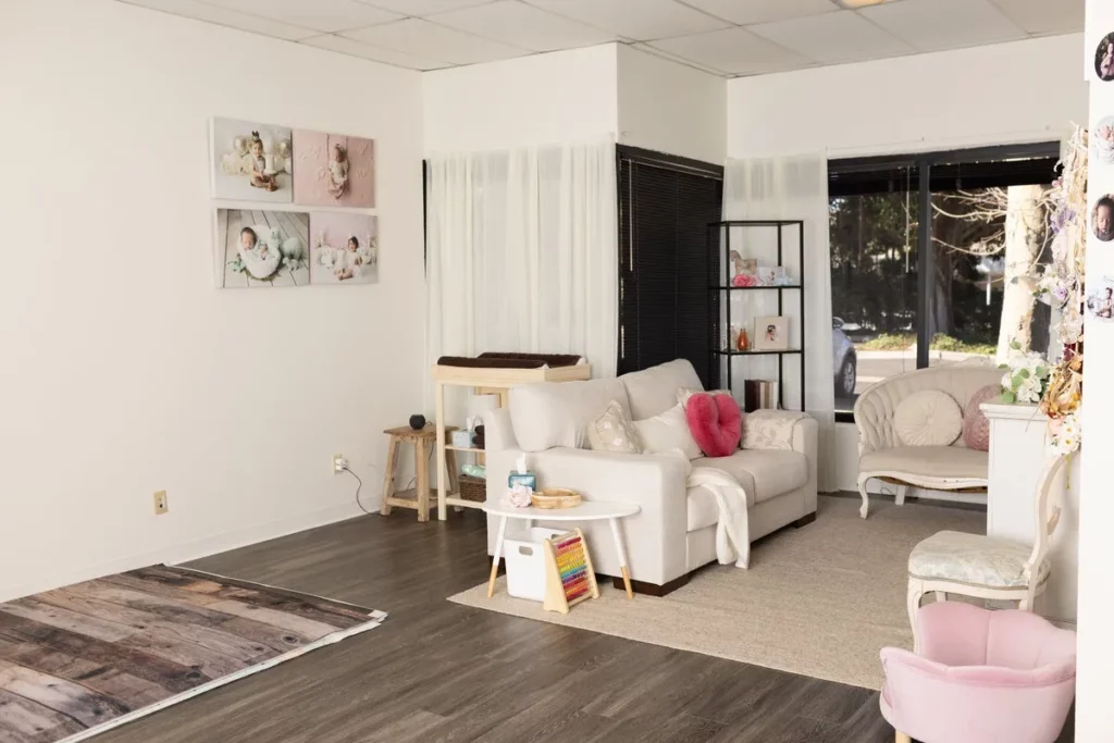 Bright room with a white sofa, armchair, photos of babies on the wall, a small play area, and large windows letting in natural light. Lovely Baby Photography by Stacey Marsh