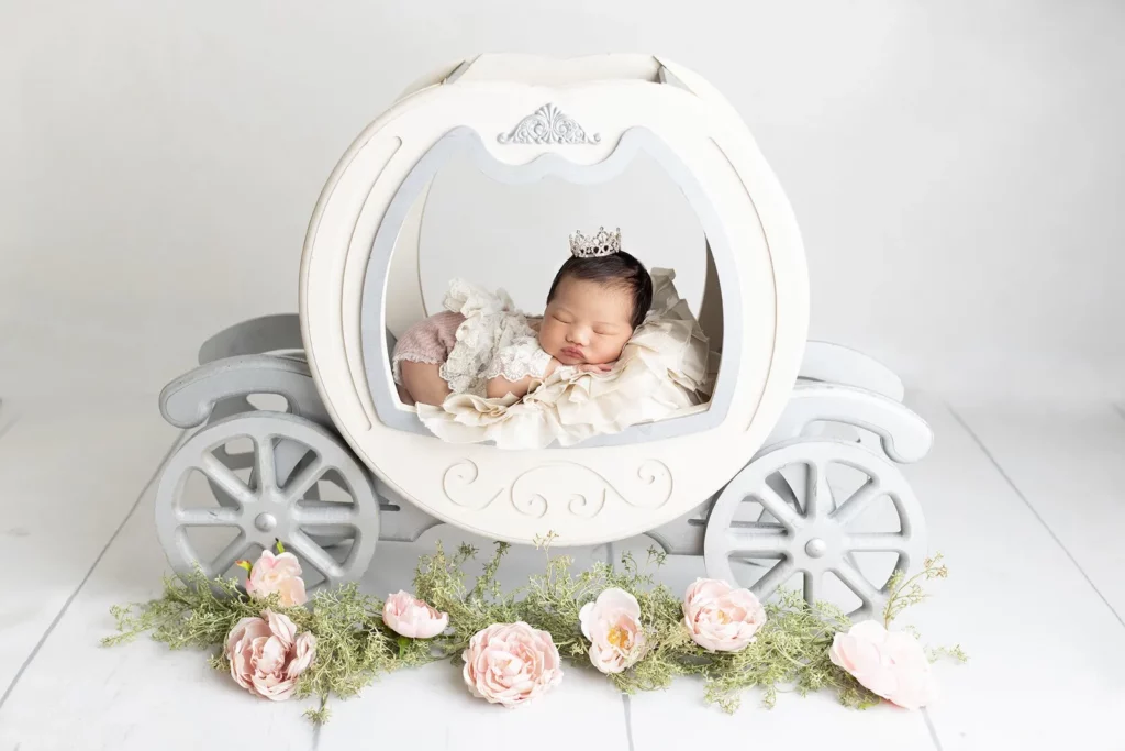 A newborn baby wearing a small crown sleeps inside a white, decorative carriage prop, surrounded by pink flowers and greenery on a white background. Lovely Baby Photography by Stacey Marsh