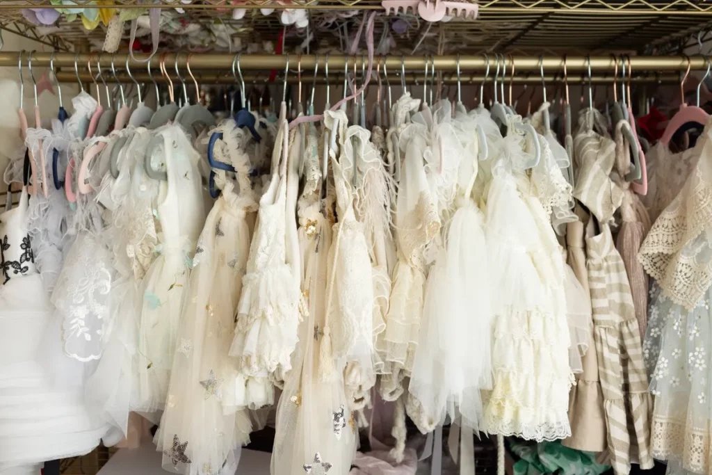 Several frilly, cream and white dresses with lace and tulle details are hanging on a clothing rack with assorted hangers. Lovely Baby Photography by Stacey Marsh