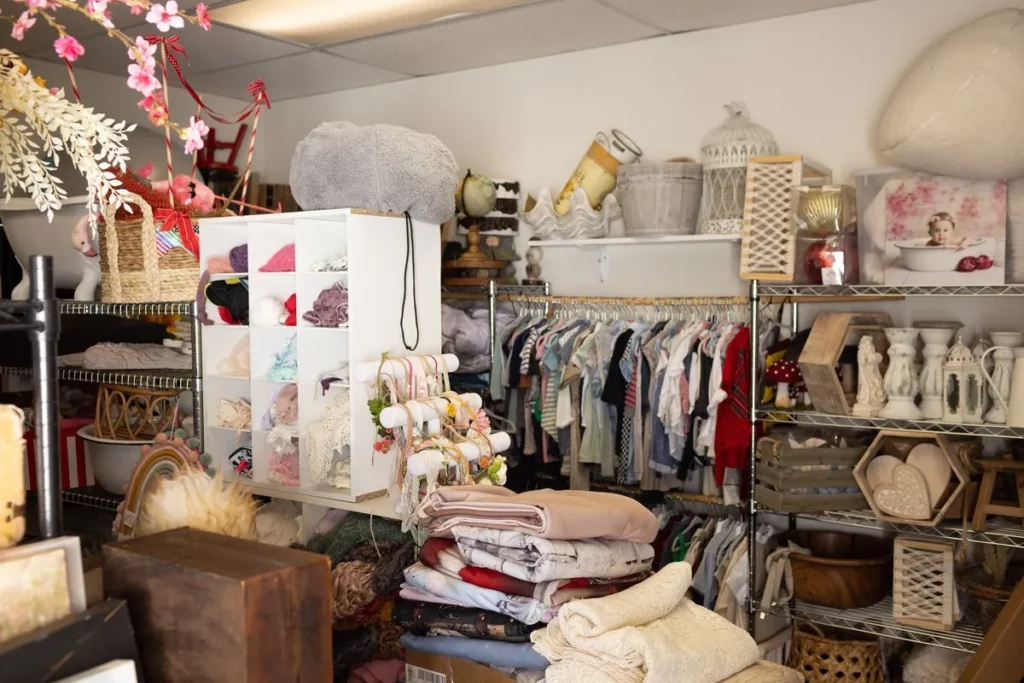 A small shop interior with shelves and racks holding clothes, folded linens, baskets, jars, decorative items, and various household goods. Lovely Baby Photography by Stacey Marsh