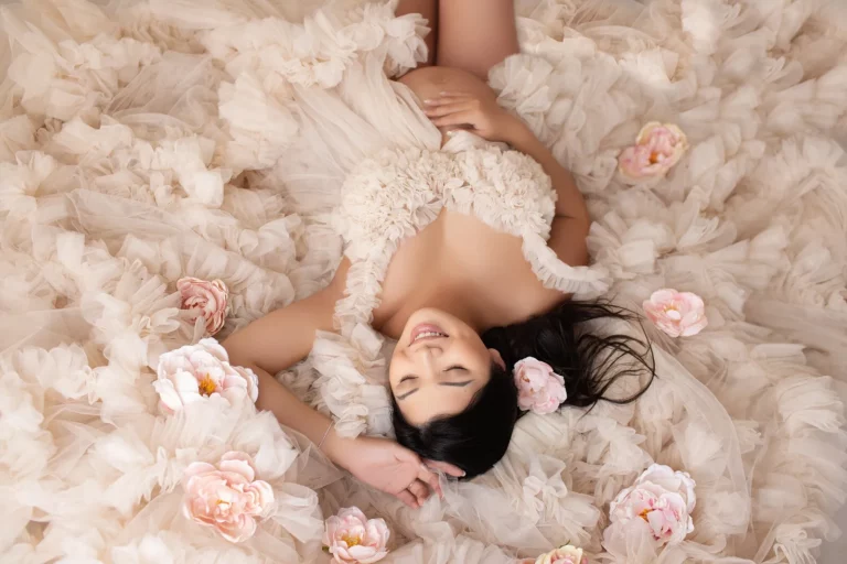 Woman in a ruffled cream dress lies on a matching floral fabric, surrounded by pale pink flowers, with eyes closed and a flower in her hair. Lovely Baby Photography by Stacey Marsh