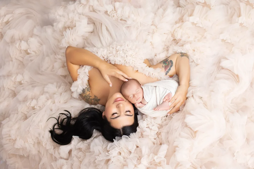 A woman in a ruffled light pink dress lies on her back, holding a swaddled newborn baby, both facing up and surrounded by soft fabric. Lovely Baby Photography by Stacey Marsh
