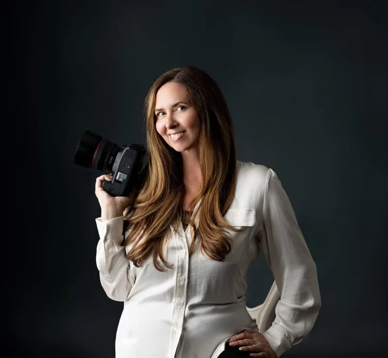 A woman with long brown hair in a white blouse holds a camera on her shoulder, standing against a dark background and smiling. Lovely Baby Photography by Stacey Marsh