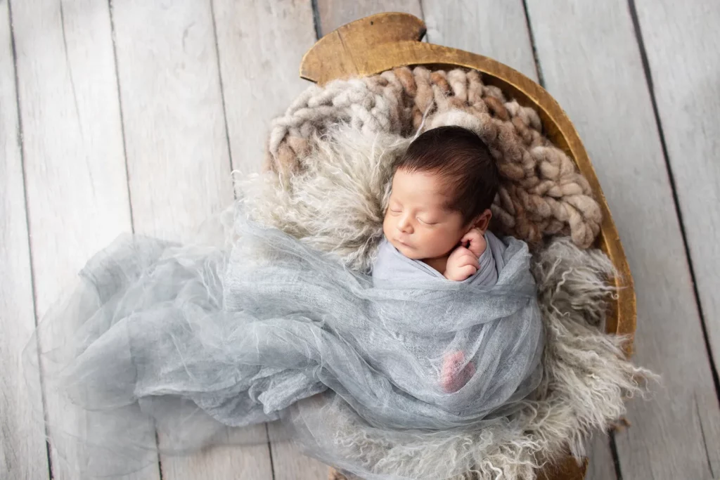 Newborn baby wrapped in a light gray swaddle, sleeping on a furry blanket in a wooden basket on a white wooden floor. Lovely Baby Photography by Stacey Marsh