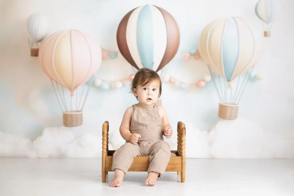 A young child sits on a small wooden bed in front of a pastel hot air balloon-themed backdrop with clouds and garlands.