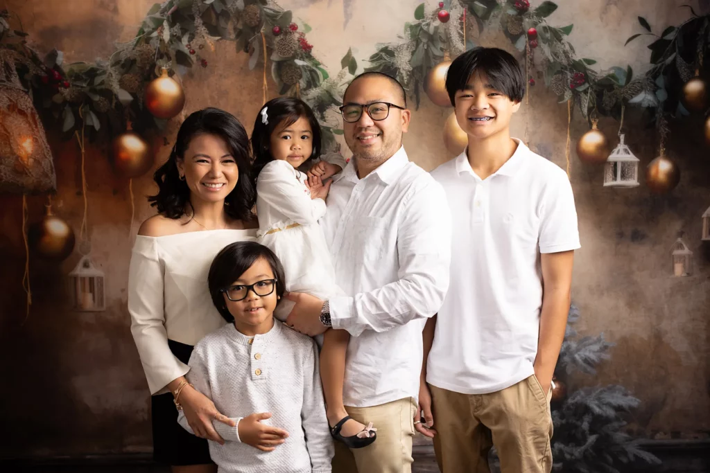 A family of five poses together in front of a festive holiday backdrop with gold ornaments and greenery, all dressed in white and beige clothing. Lovely Baby Photography, 2185 Faraday Ave suite 110, Carlsbad, CA 92008, +1 760-271-0725