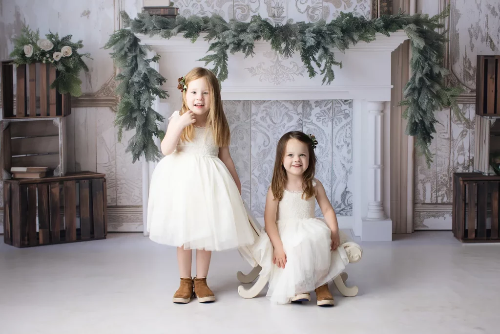 Two young girls in white dresses and brown boots pose in front of a decorated white mantel with greenery, one standing and one sitting on a small sled. Lovely Baby Photography, 2185 Faraday Ave suite 110, Carlsbad, CA 92008, +1 760-271-0725