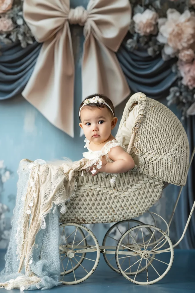 A baby wearing a headband and lace outfit sits in a vintage wicker stroller, with a backdrop of flowers and large fabric bows.