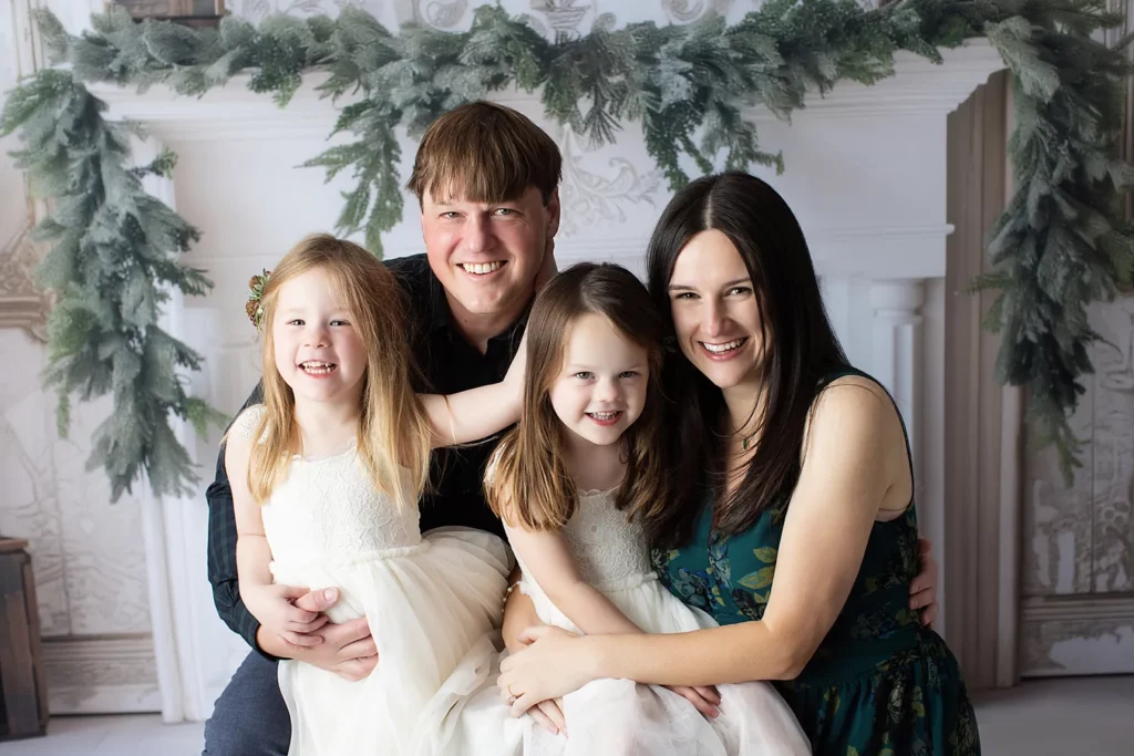 A man and woman sit closely together, each holding a young girl in white dresses, with greenery and a white mantle in the background. All four are smiling at the camera. Lovely Baby Photography, 2185 Faraday Ave suite 110, Carlsbad, CA 92008, +1 760-271-0725