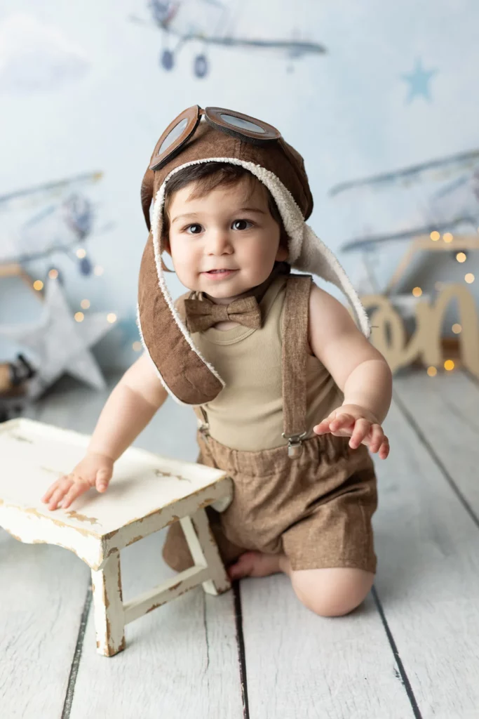 A baby wearing aviator goggles and brown overalls kneels on a wooden floor next to a small white bench, with a backdrop featuring airplane illustrations.