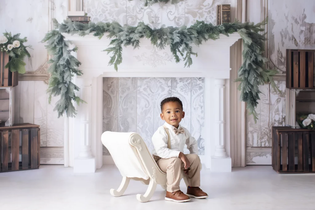 A young boy in dress clothes sits on a small white bench in front of a decorated white fireplace with greenery garland. Lovely Baby Photography, 2185 Faraday Ave suite 110, Carlsbad, CA 92008, +1 760-271-0725