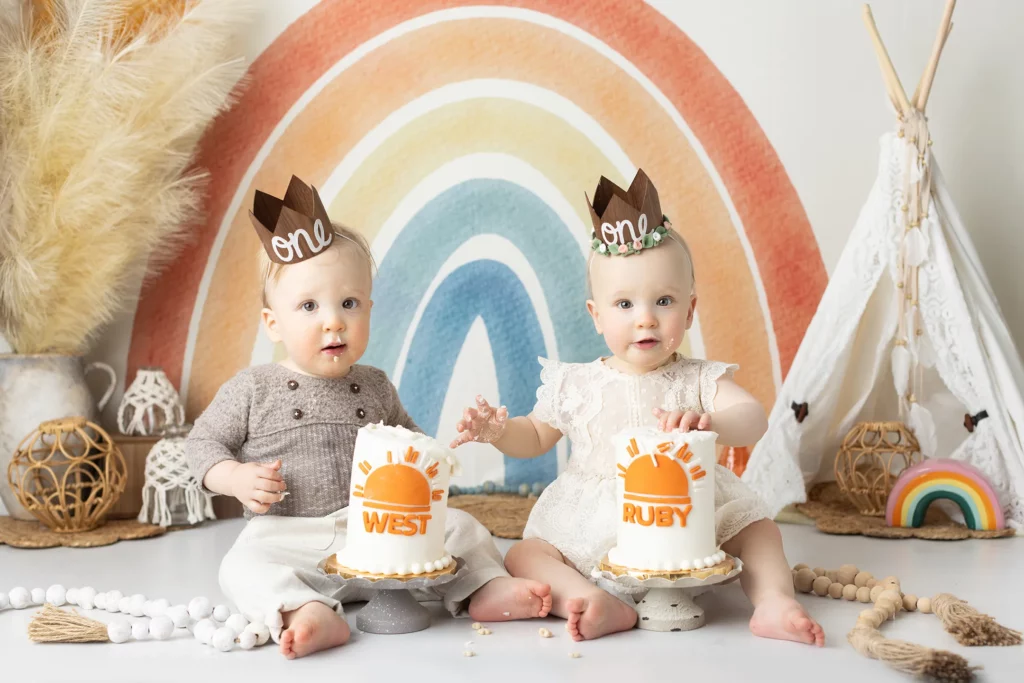 Two babies wearing "one" crowns sit in front of rainbow decor with cakes labeled "WEST" and "RUBY," surrounded by boho-style decorations.