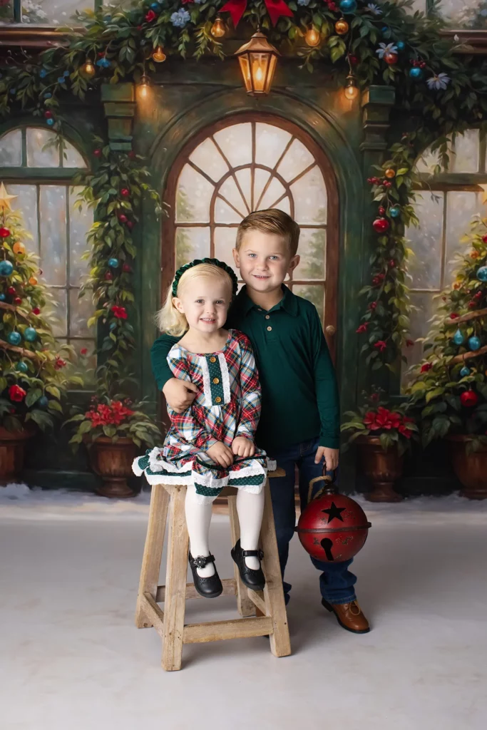 Two young children pose for a holiday photo; the girl sits on a stool in a plaid dress, while the boy stands beside her holding a large red jingle bell. Festive backdrop with greenery and lights. Lovely Baby Photography, 2185 Faraday Ave suite 110, Carlsbad, CA 92008, +1 760-271-0725