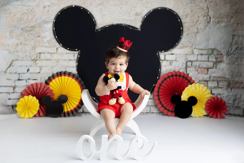 Baby wearing a red outfit and crown sits on a white chair holding a Mickey Mouse toy, with a "one" sign and Mickey-themed decorations in the background.