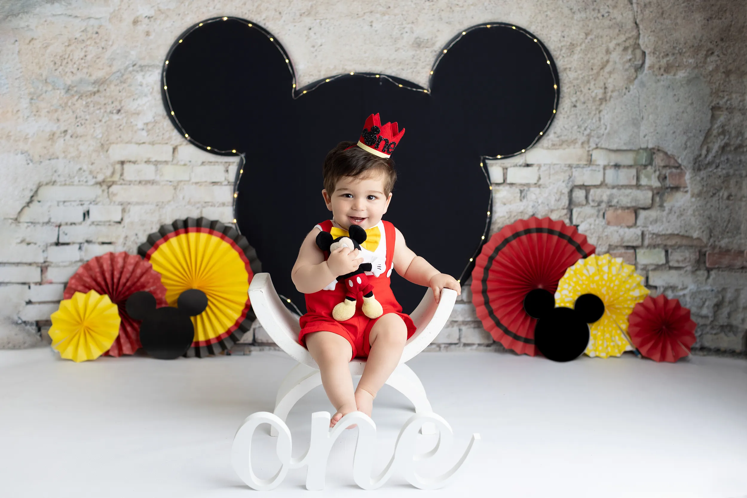 Baby wearing a red outfit and crown sits on a white chair holding a Mickey Mouse toy, with a "one" sign and Mickey-themed decorations in the background. Lovely Baby Photography, 2185 Faraday Ave suite 110, Carlsbad, CA 92008, +1 760-271-0725