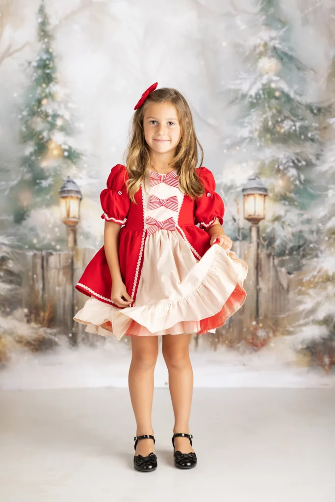 Young girl stands indoors in front of a winter-themed backdrop, wearing a red and cream dress with bows and black shoes, holding up the edge of her skirt. Lovely Baby Photography, 2185 Faraday Ave suite 110, Carlsbad, CA 92008, +1 760-271-0725