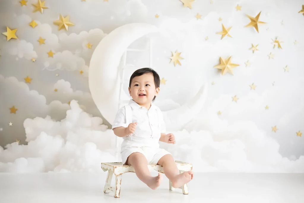A baby wearing a white outfit sits on a small bench in front of a backdrop with clouds, a crescent moon, and gold stars.
