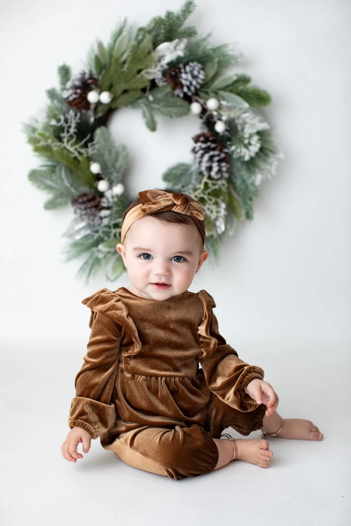 Baby wearing a brown velvet outfit and headband sits in front of a festive green wreath with pine cones and white accents. Lovely Baby Photography, 2185 Faraday Ave suite 110, Carlsbad, CA 92008, +1 760-271-0725