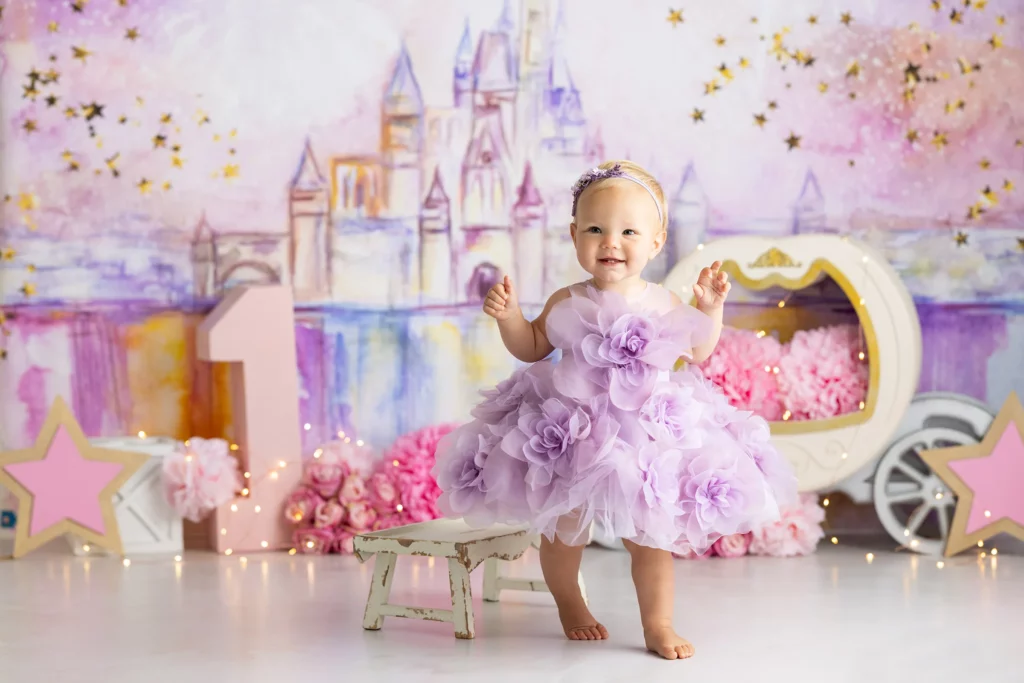 A baby in a lavender floral dress stands smiling in front of a pastel castle backdrop, with pink decorations and a large number one, suggesting a first birthday celebration.