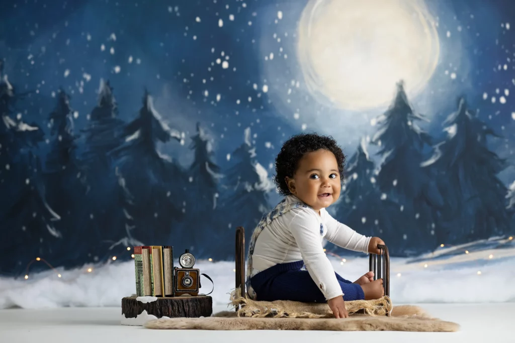 A baby sits on a fur rug with a small sled, surrounded by books and a vintage camera, against a painted snowy forest and full moon backdrop. Lovely Baby Photography, 2185 Faraday Ave suite 110, Carlsbad, CA 92008, +1 760-271-0725