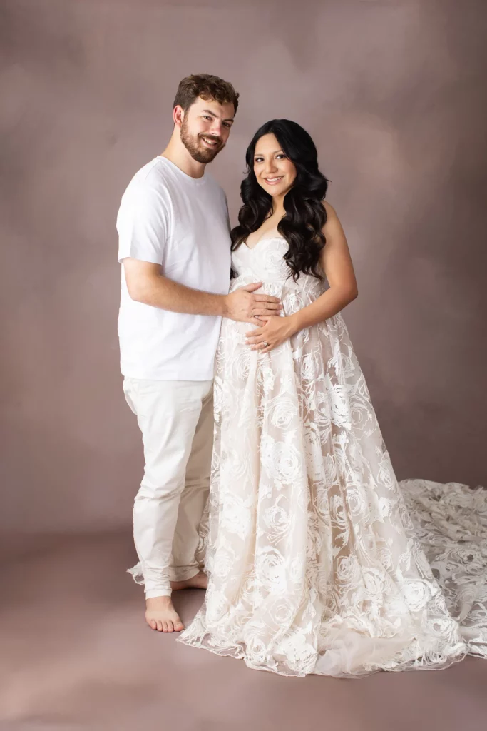 A man in a white shirt and pants stands next to a pregnant woman in a white lace dress, both smiling and posing against a neutral studio backdrop.