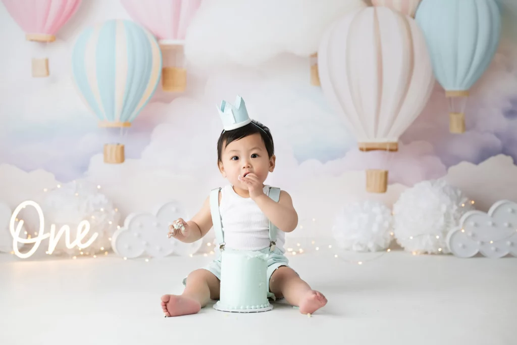 A baby wearing a crown sits on the floor, eating cake, with a pastel hot air balloon backdrop and the word "One" illuminated beside them.