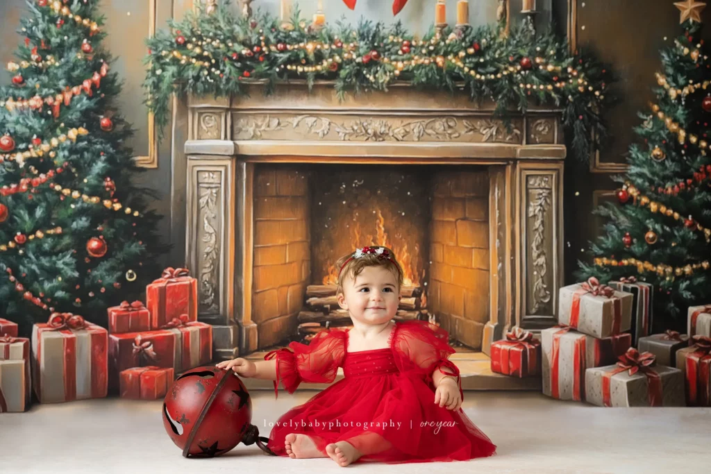 A baby in a red dress sits on the floor in front of a decorated Christmas fireplace, surrounded by wrapped gifts and holding a red ornament. Lovely Baby Photography, 2185 Faraday Ave suite 110, Carlsbad, CA 92008, +1 760-271-0725