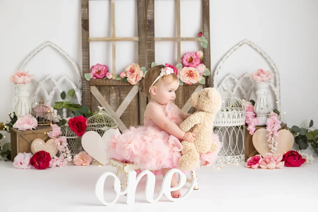 A baby in a pink dress sits holding a teddy bear, surrounded by floral decorations and vintage props, with a large “one” sign in front.