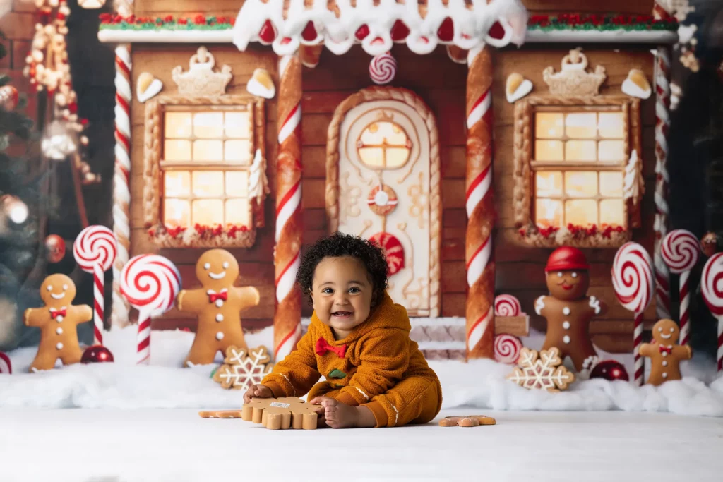 A baby in a brown reindeer outfit sits on the floor with gingerbread cookies, in front of a gingerbread house decorated with candy canes and gingerbread figures. Lovely Baby Photography, 2185 Faraday Ave suite 110, Carlsbad, CA 92008, +1 760-271-0725