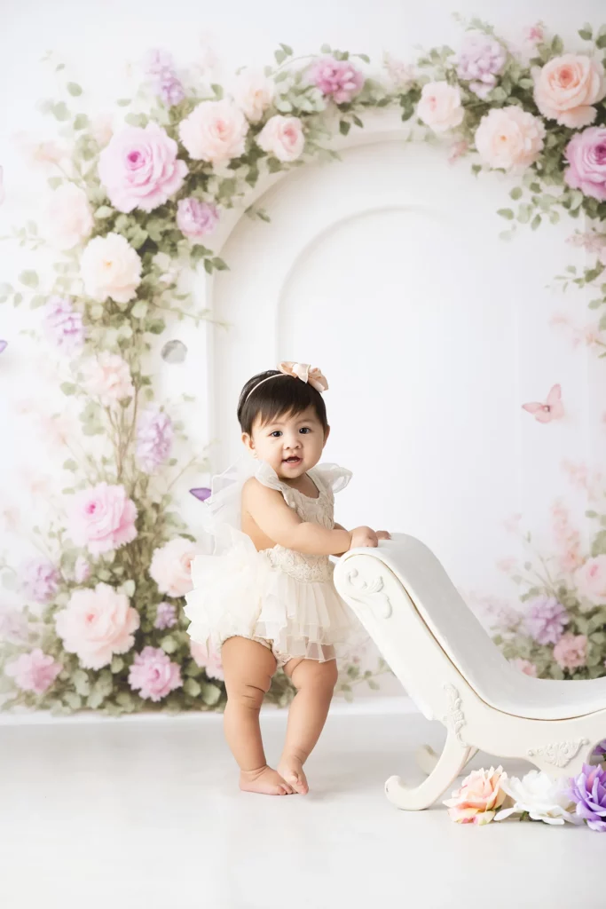 A baby in a frilly cream outfit stands next to a white chaise lounge, with a floral arch of pink and purple flowers in the background.