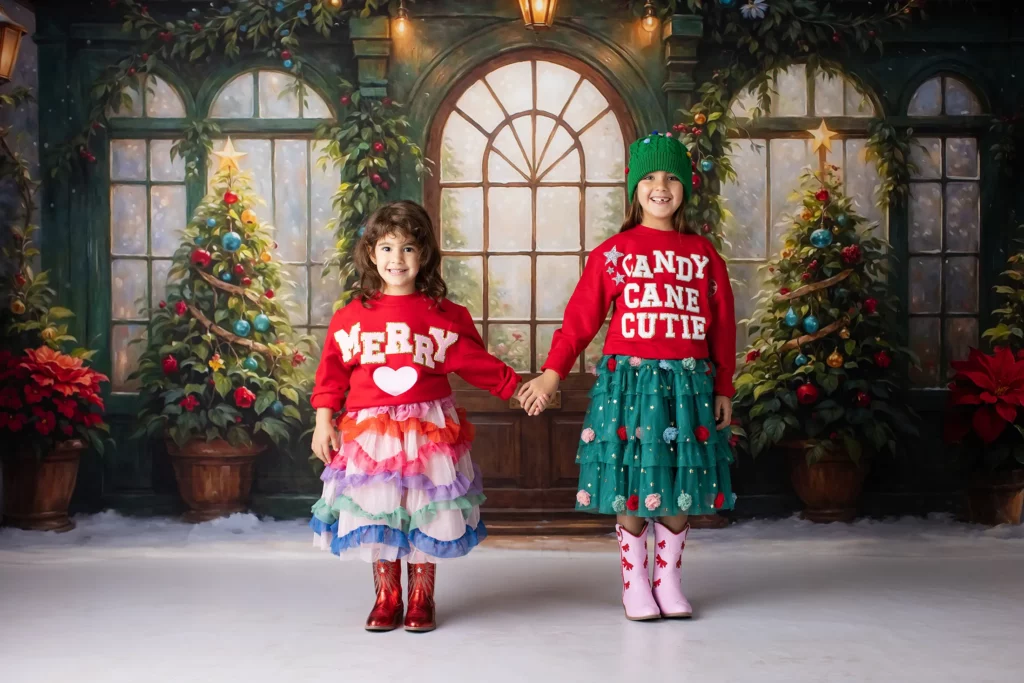Two children stand holding hands in festive outfits in front of a Christmas-themed backdrop with decorated trees and poinsettias. Lovely Baby Photography, 2185 Faraday Ave suite 110, Carlsbad, CA 92008, +1 760-271-0725