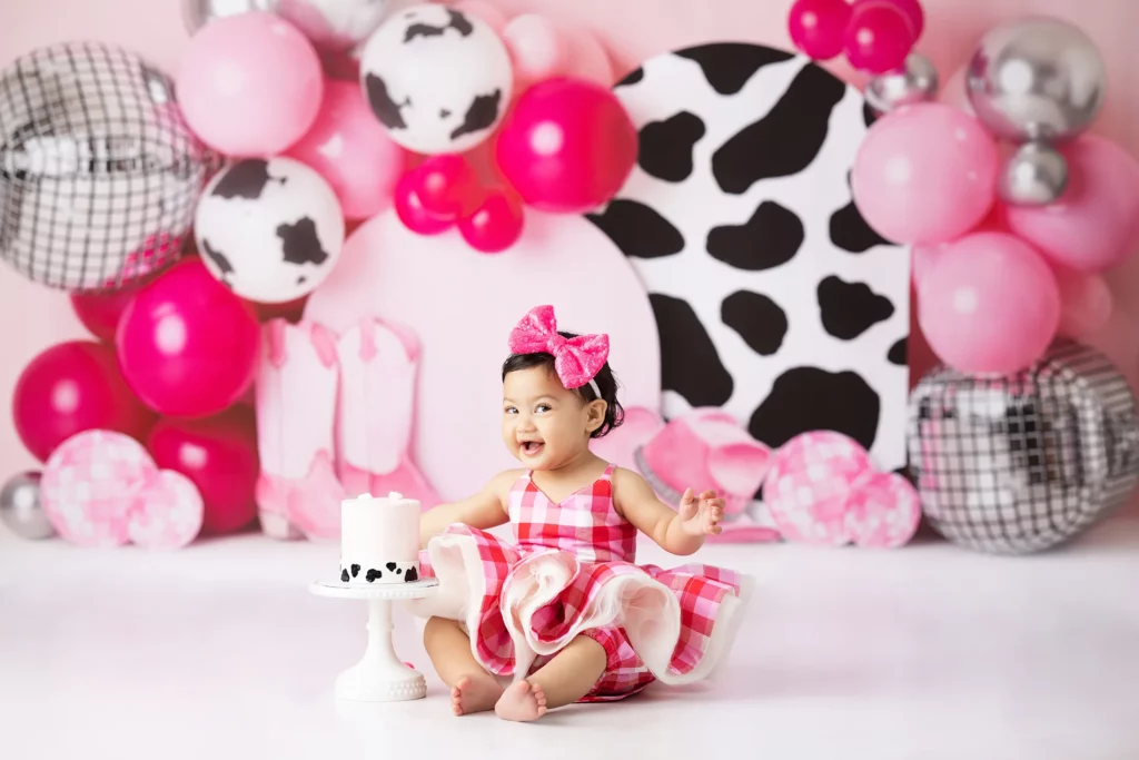 A young child in a pink plaid dress sits on the floor with a small cake, surrounded by pink and cow-patterned balloons and decorations.
