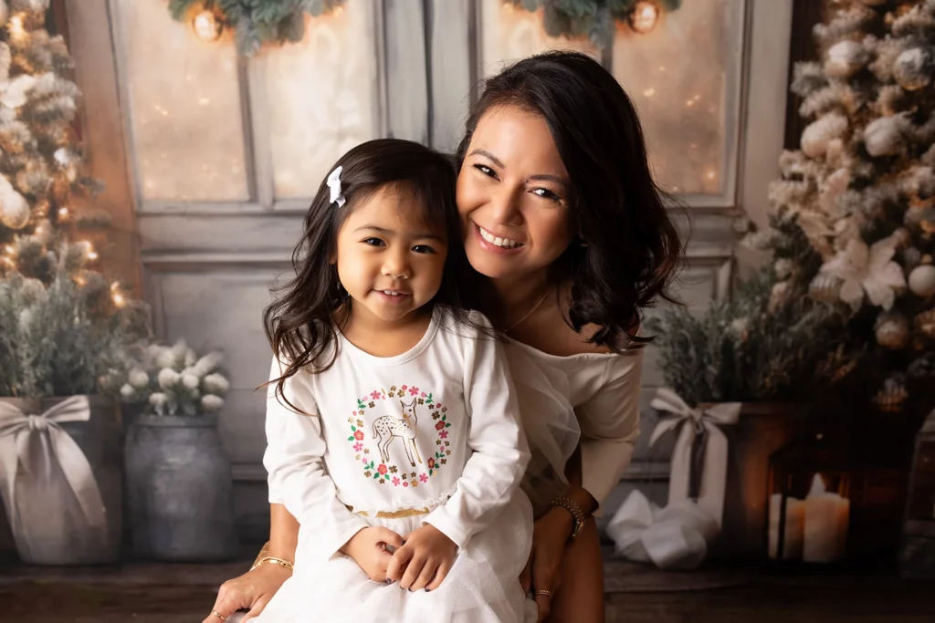 A woman and a young girl dressed in white pose together, smiling, in front of a festive, winter-themed backdrop with decorated trees and soft lighting. Lovely Baby Photography, 2185 Faraday Ave suite 110, Carlsbad, CA 92008, +1 760-271-0725