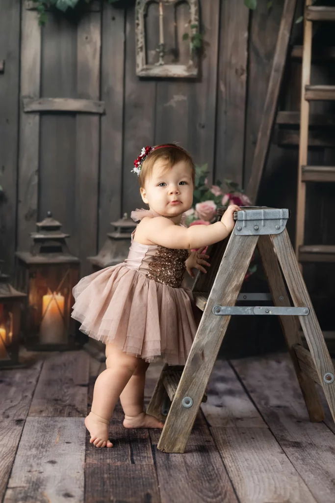 A toddler in a brown dress stands holding a small wooden ladder, with lanterns and flowers in the background on a wooden floor.