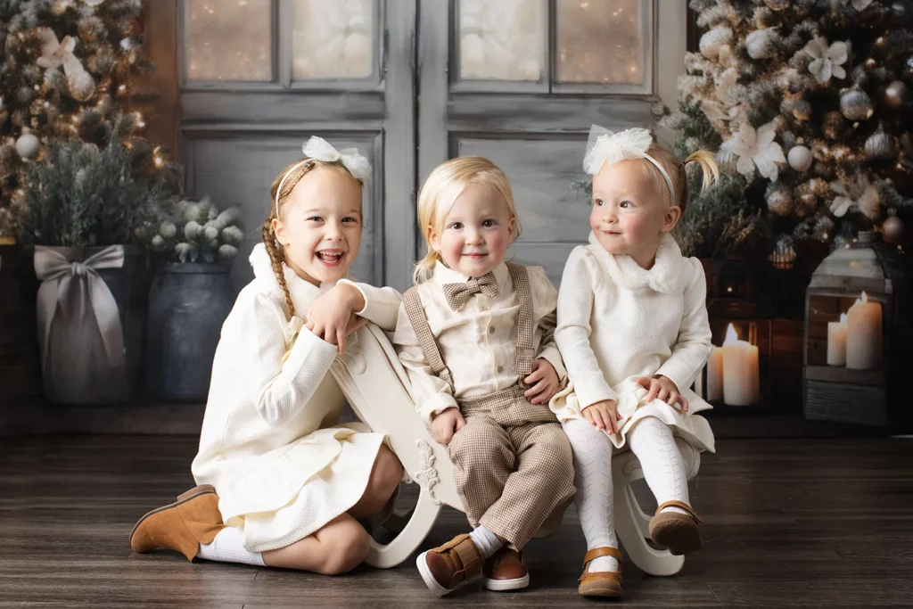 Three young children in festive winter outfits sit and pose in front of decorated Christmas trees, candles, and a wooden door backdrop. Lovely Baby Photography, 2185 Faraday Ave suite 110, Carlsbad, CA 92008, +1 760-271-0725