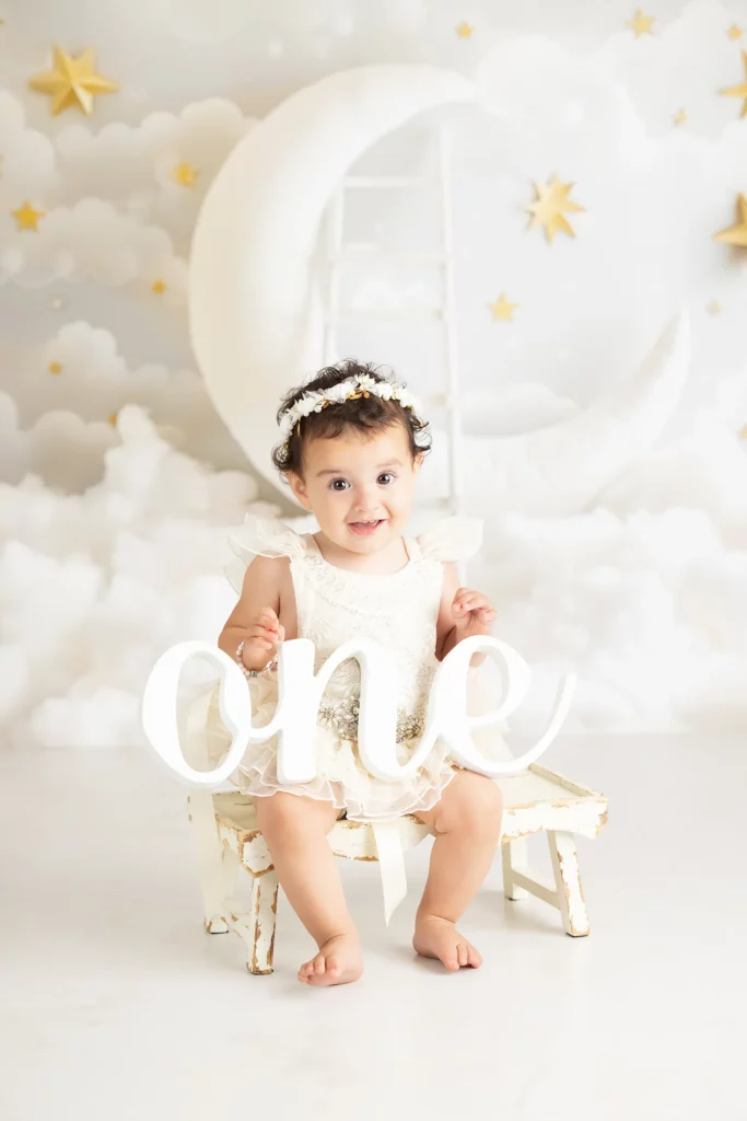 A baby girl in a white dress and floral headband sits on a small bench, holding a sign that says “one,” with a cloud and moon backdrop decorated with stars.