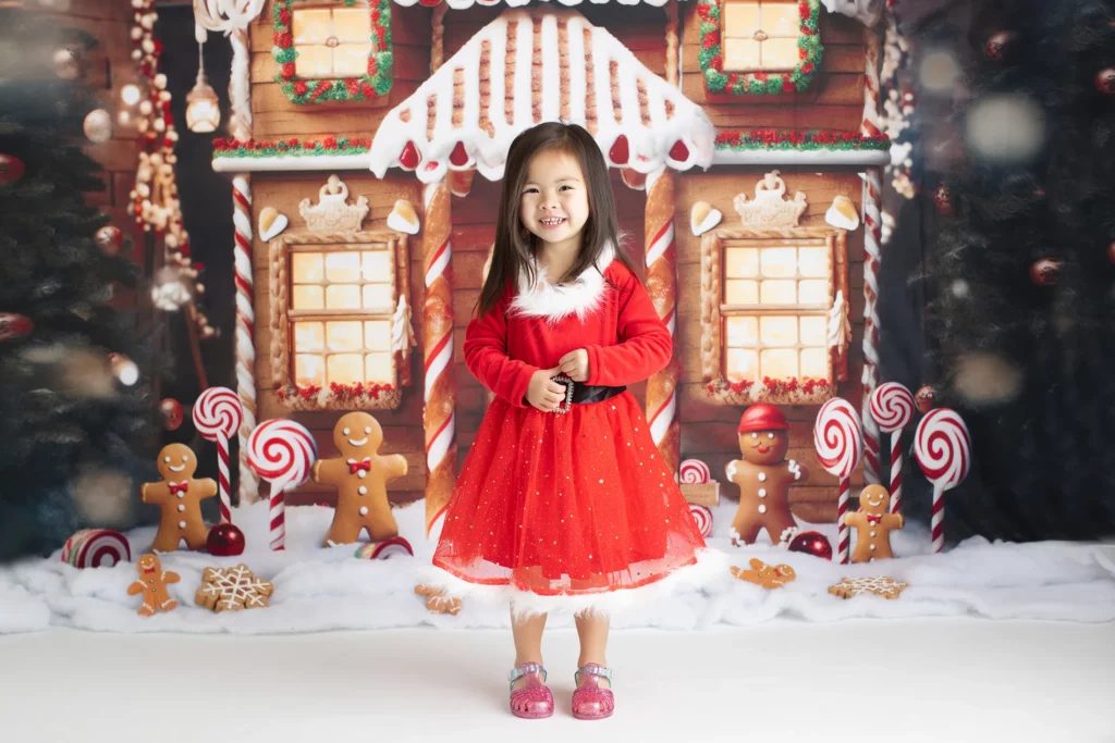 A young girl in a red holiday dress stands in front of a festive gingerbread house backdrop decorated with candy canes and gingerbread figures. Lovely Baby Photography, 2185 Faraday Ave suite 110, Carlsbad, CA 92008, +1 760-271-0725