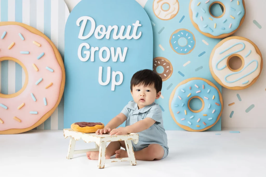 A young child sits on the floor in front of a small table with a donut, surrounded by large donut decorations and a sign that says "Donut Grow Up.