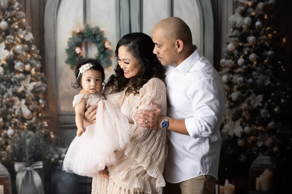 A man and woman stand together, holding a young girl in a pink dress. Behind them are decorated Christmas trees, wreaths, and candles. Lovely Baby Photography, 2185 Faraday Ave suite 110, Carlsbad, CA 92008, +1 760-271-0725