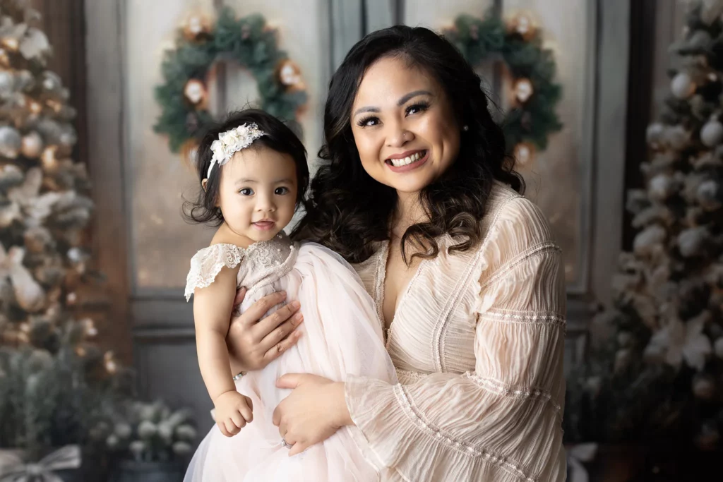 A woman in a light dress holds a young girl in a pale dress and headband, posing in front of a festive backdrop with holiday wreaths and decorated trees. Lovely Baby Photography, 2185 Faraday Ave suite 110, Carlsbad, CA 92008, +1 760-271-0725