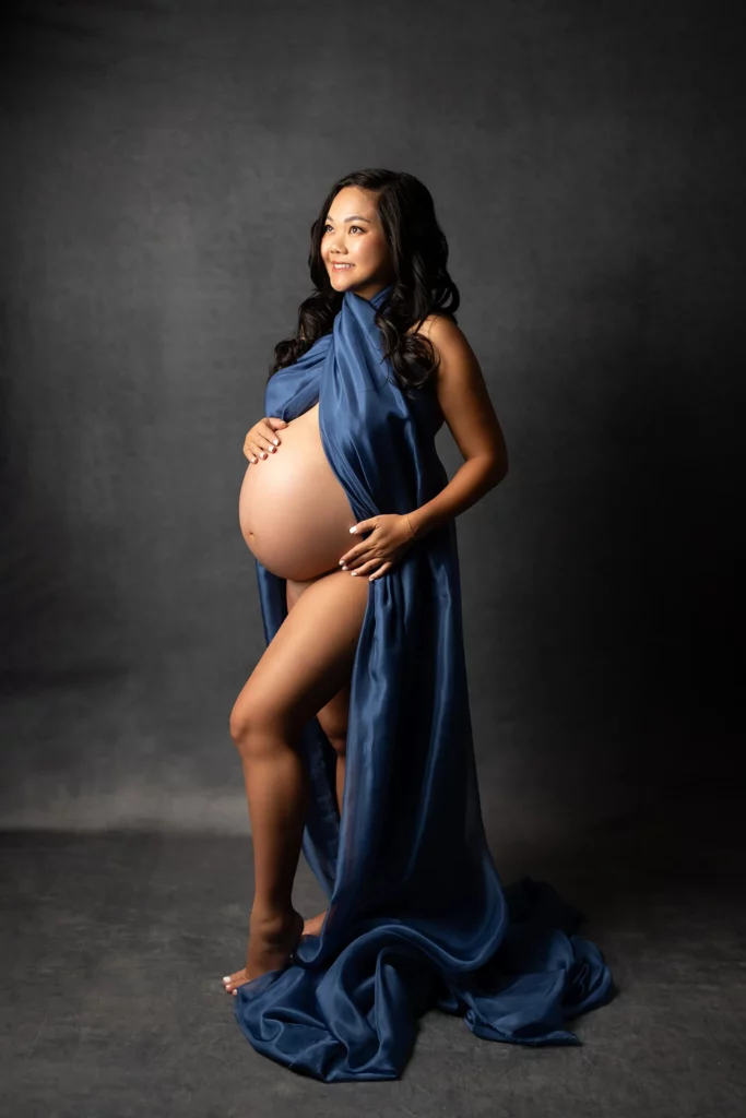 A pregnant woman stands barefoot, draped in a blue satin fabric, holding her belly and smiling against a dark, plain background.