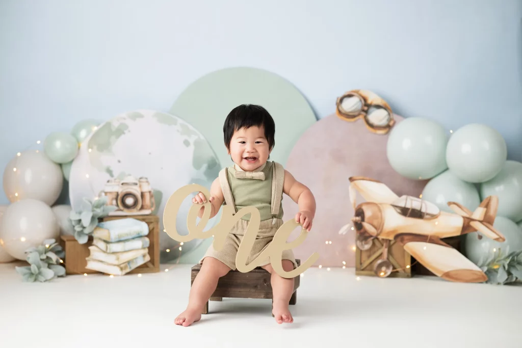 A smiling baby in a green outfit sits on a wooden bench holding a "one" sign, surrounded by vintage travel-themed decorations and light blue balloons.