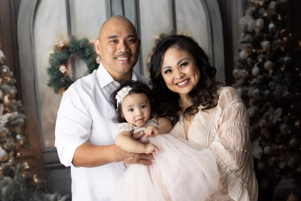 A man, woman, and baby pose together in front of a festive, holiday-themed background with wreaths and decorated trees. Lovely Baby Photography, 2185 Faraday Ave suite 110, Carlsbad, CA 92008, +1 760-271-0725