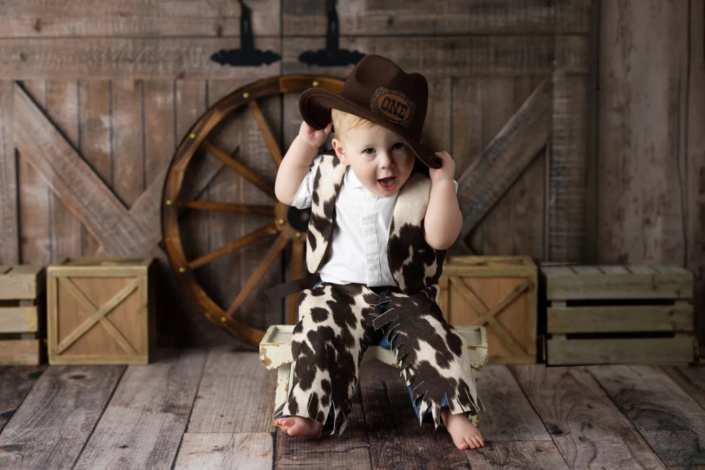 A young child in a cow-print cowboy outfit sits barefoot on a wooden bench, smiling and holding a brown cowboy hat in front of a rustic barn backdrop.